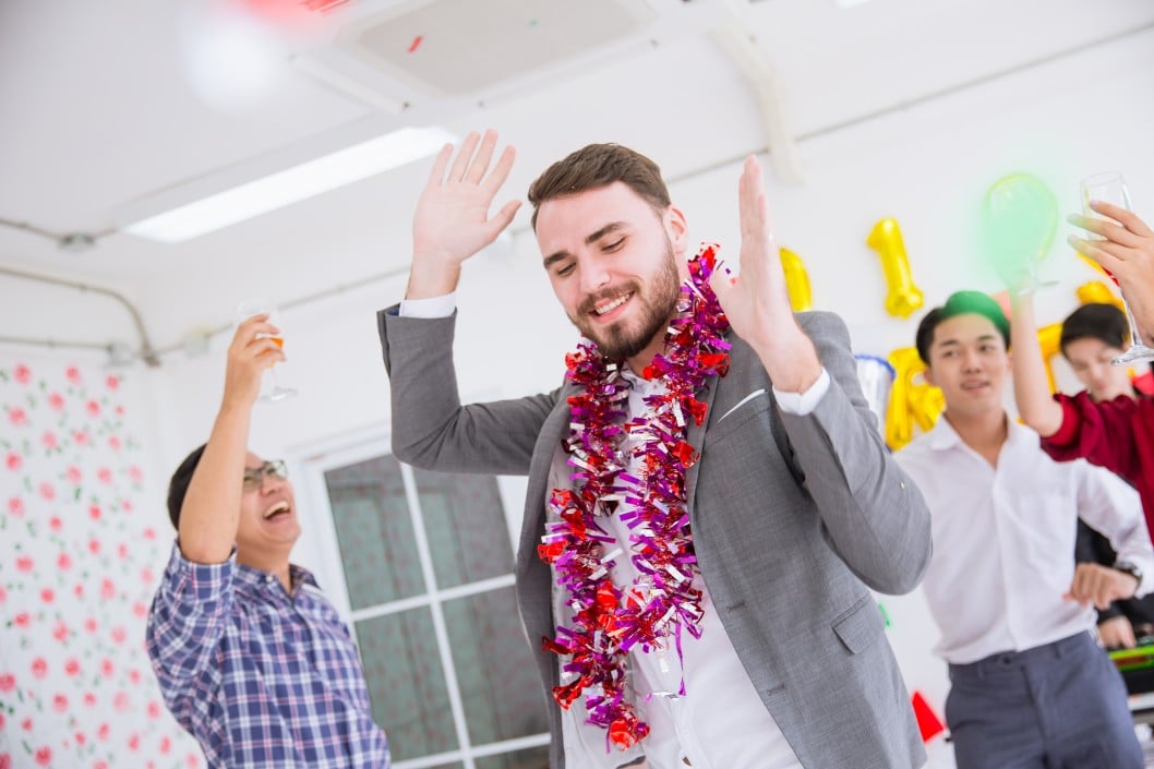 Group of festive colleagues celebrating at a corporate party, man in a suit wearing a colorful garland, raising hands joyfully, surrounded by friends holding drinks and enjoying the moment.