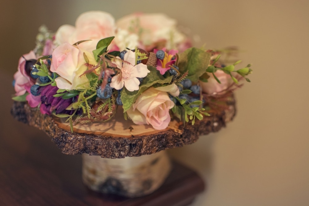 Floral arrangement featuring pink roses, purple blooms, and greenery displayed on a rustic wooden base, ideal for a winter wedding theme.