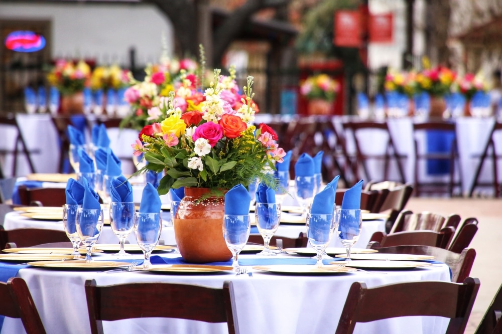 Beautifully arranged outdoor wedding table setting with colorful floral centerpieces, blue napkins, glassware, and white tablecloths, emphasizing event planning and venue aesthetics.