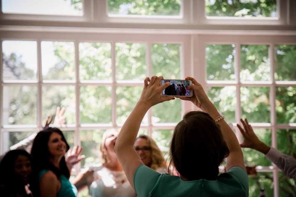 Woman taking a photo of a bridal party with a smartphone, surrounded by guests celebrating in a well-lit venue with large windows.