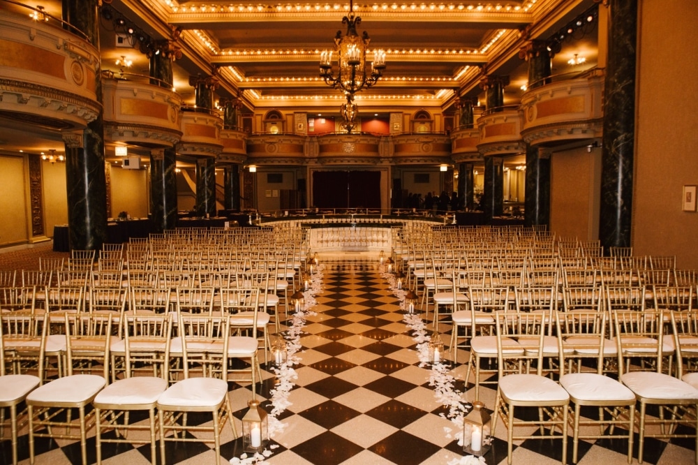 Elegant ballroom setup for a wedding ceremony, featuring rows of gold chiavari chairs, a checkered floor, floral decorations, and a chandelier above, reflecting a formal event atmosphere.