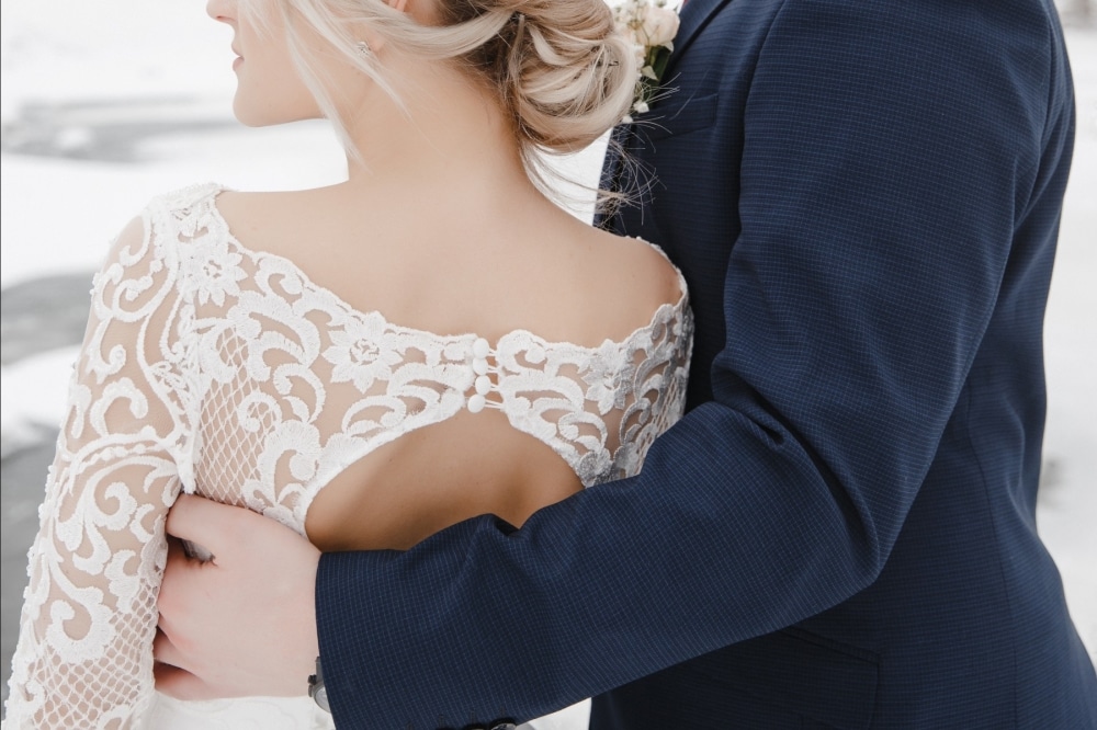 Beautiful bride in lace wedding dress with intricate back detail, embraced by groom in navy suit, set against a snowy landscape, symbolizing love and winter wedding elegance.