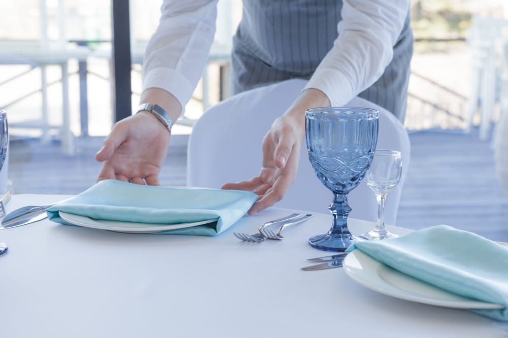 Waiter setting a wedding table with blue napkins, plates, and glassware, emphasizing event preparation and dining arrangements.