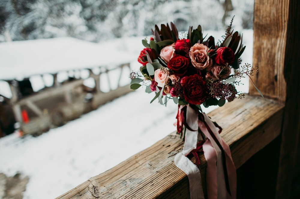 Winter wedding bouquet featuring deep burgundy and plum roses, blush accents, and greenery, set against a snowy outdoor backdrop.