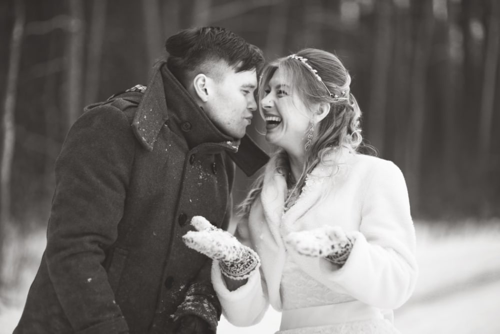 Newly married couple laughing and playing in snow, wearing winter wedding attire, in a snowy forest setting, evoking a romantic and joyful winter wedding theme.