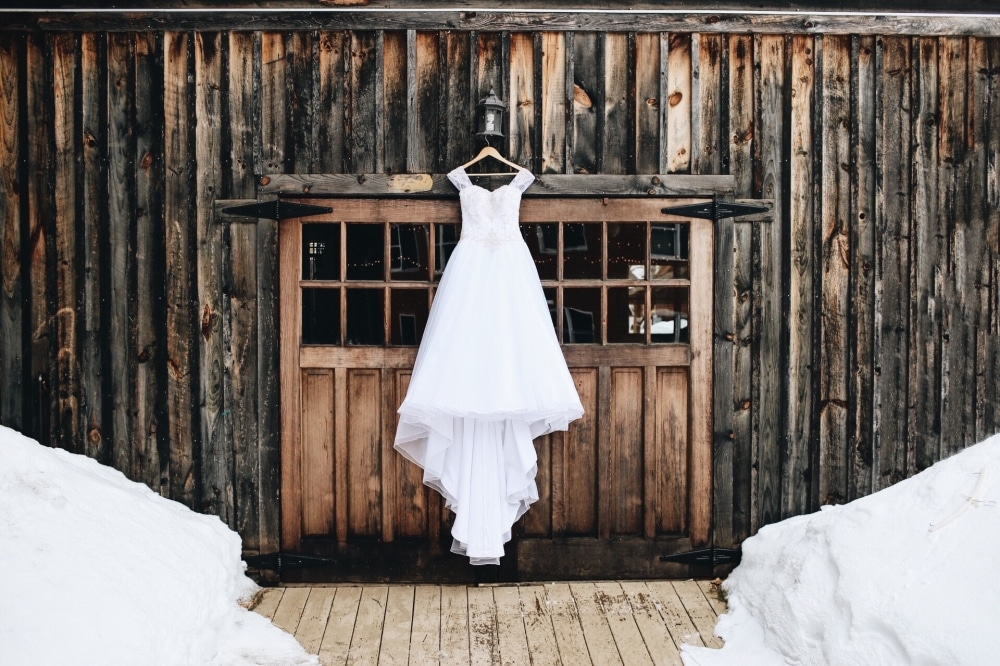 Wedding dress hanging on a wooden barn door, surrounded by snow, representing a winter farmhouse wedding theme.