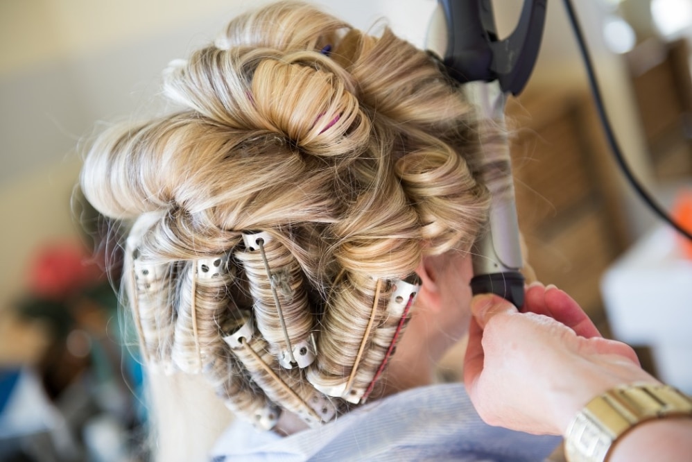 Blonde hair being styled with curling rollers and a curling iron, showcasing a hairstylist preparing for a wedding hairstyle.
