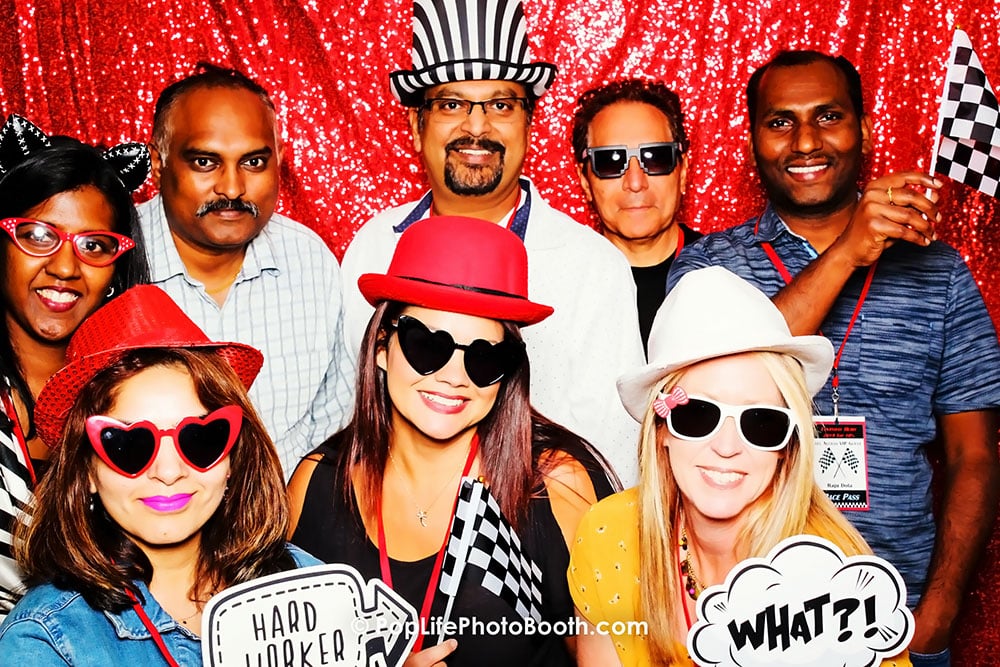 Group of diverse individuals in playful attire posing in a photo booth, featuring fun props and a sparkling red backdrop, capturing a joyful moment at an event.