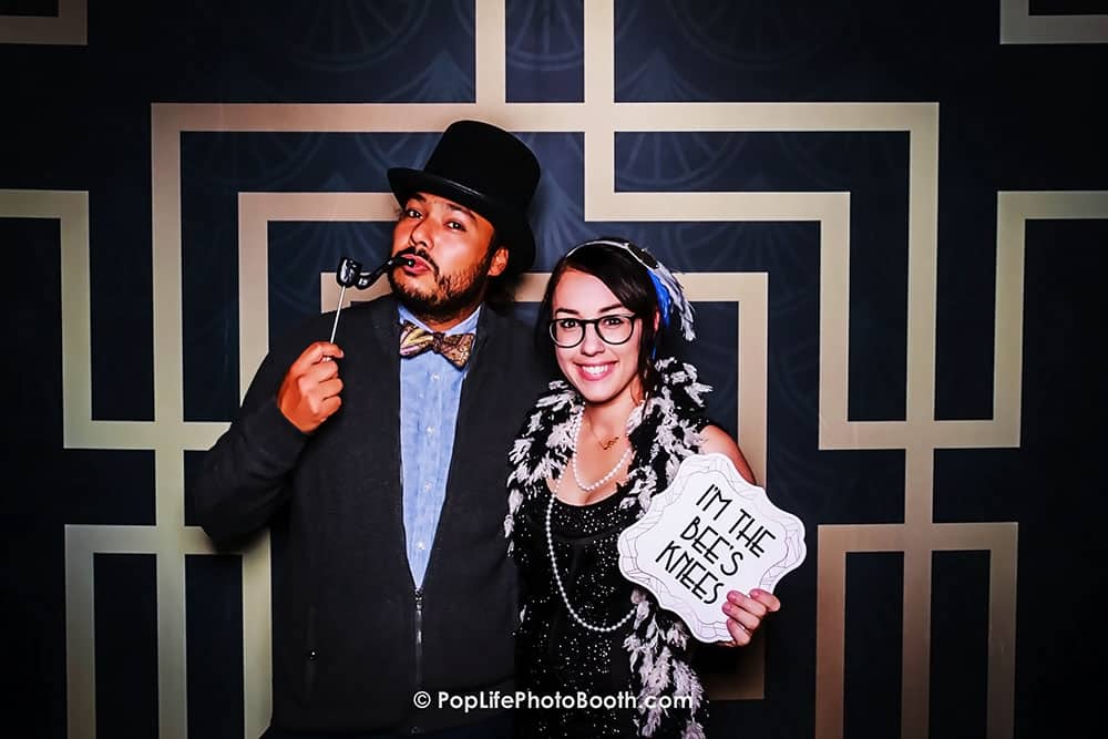 Couple posing in a photo booth with playful props, including a sign reading "I'M THE BEES KNEES," highlighting fun and memorable moments at events.