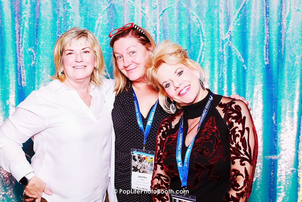 Three women smiling together in a photo booth, wearing event lanyards, against a colorful backdrop, showcasing the fun and vibrant atmosphere of events enhanced by Pop Life Photo Booth services.