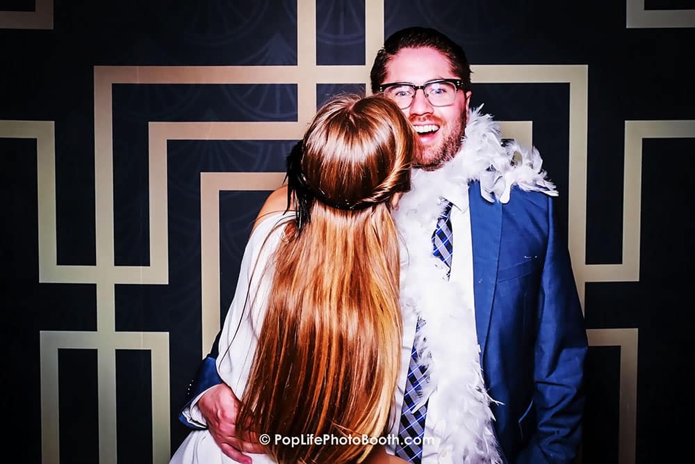 Happy couple enjoying a fun moment in a photo booth, wearing festive attire and a feather boa, against a stylish patterned backdrop, emphasizing celebration and joy at a private event.