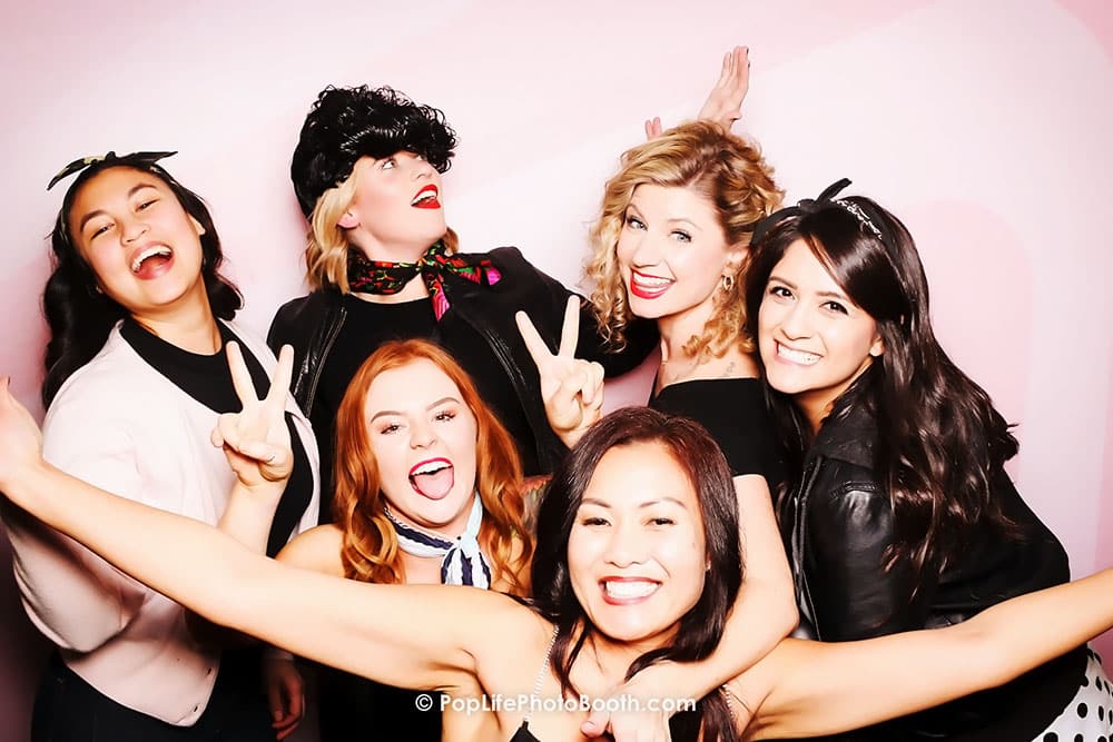 Group of six smiling women posing playfully in a photo booth, showcasing joy and fun during a private event, with a pink backdrop and stylish outfits.