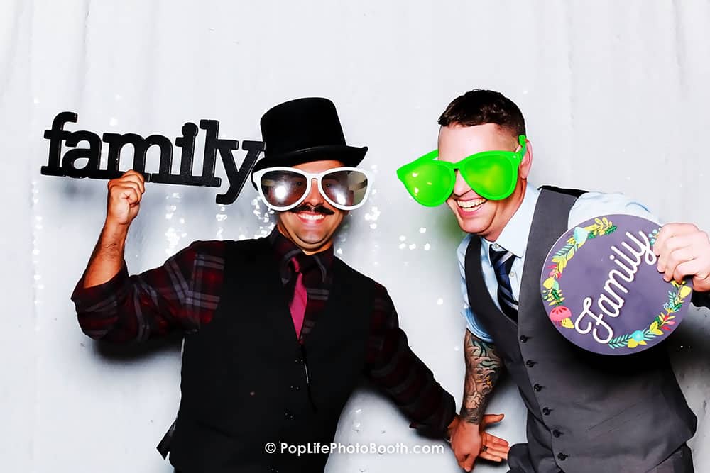 Two men wearing oversized novelty glasses and holding signs that say "family," celebrating in a photo booth with a festive backdrop, capturing joyful moments at an event.