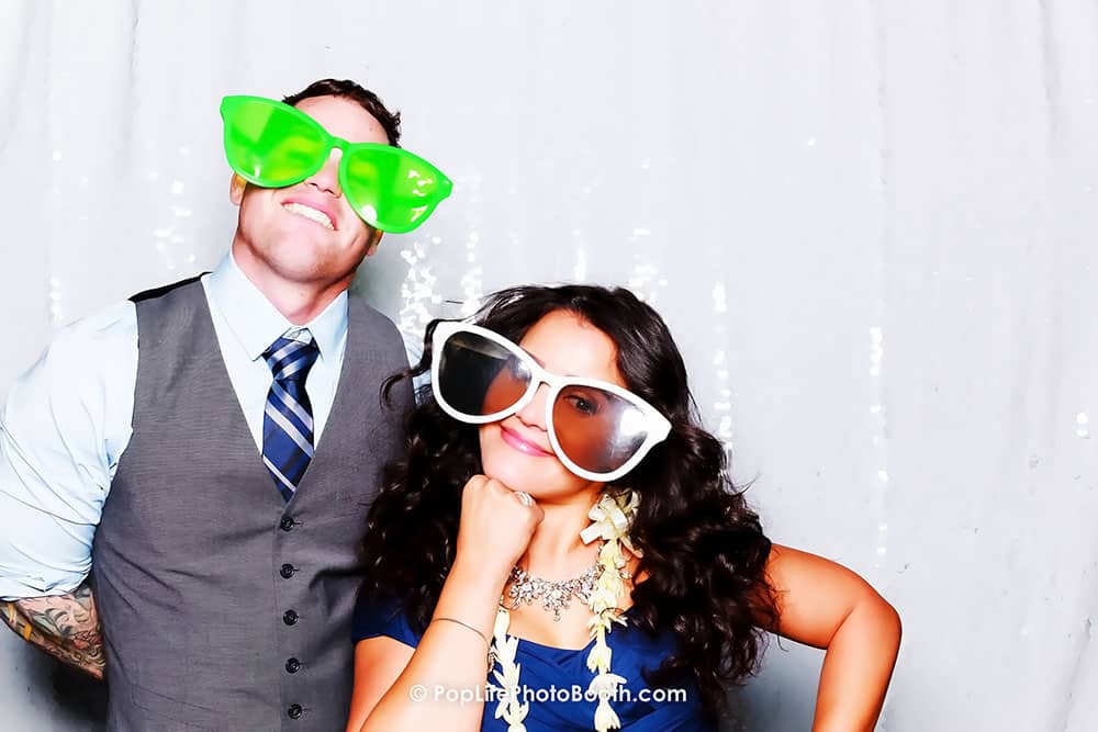 Cheerful couple posing in a photo booth, wearing oversized novelty glasses, capturing fun memories at a wedding celebration.