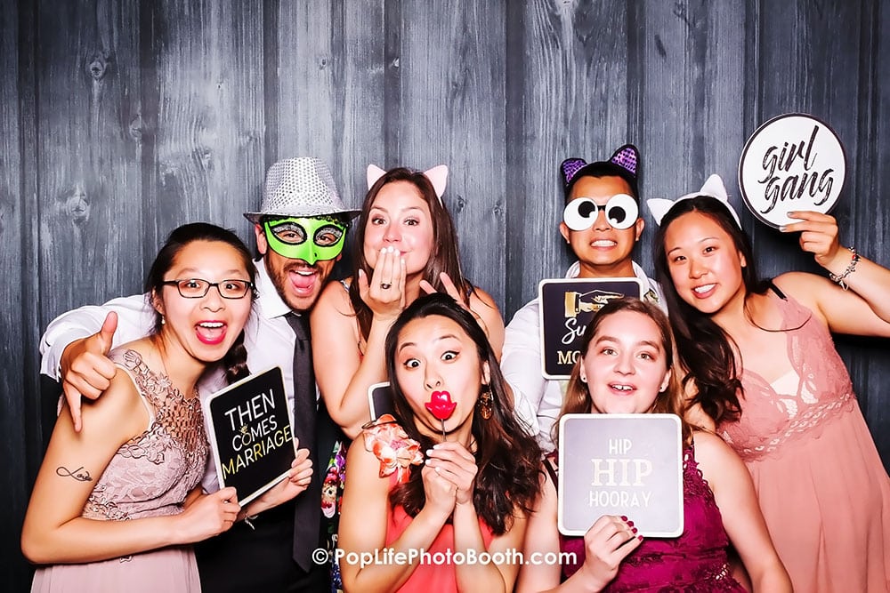 Group of joyful guests posing in a photo booth with fun props and signs, celebrating a wedding event, showcasing vibrant expressions and playful atmosphere.