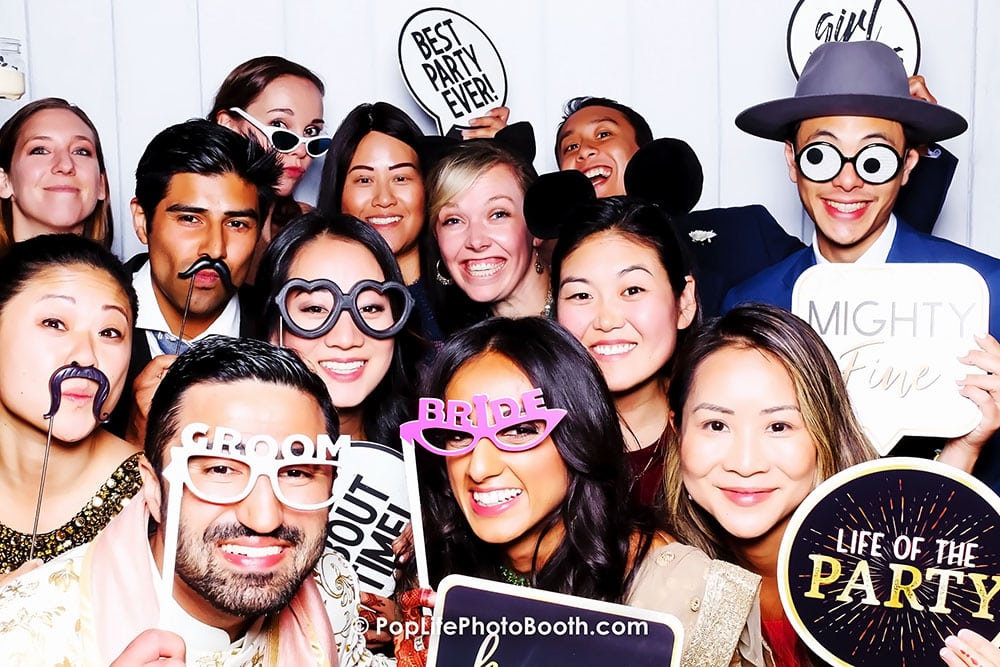 Group of joyful guests at a wedding photo booth, wearing fun props like glasses and mustaches, holding signs saying "GROOM," "BRIDE," and "BEST PARTY EVER," capturing lively moments and creating lasting memories.