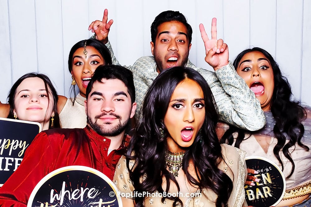 Group of friends celebrating at a wedding photo booth, showcasing joyful expressions and fun props, with signs reading "Happy Life" and "Where the Fun Begins," emphasizing the lively atmosphere of wedding festivities.