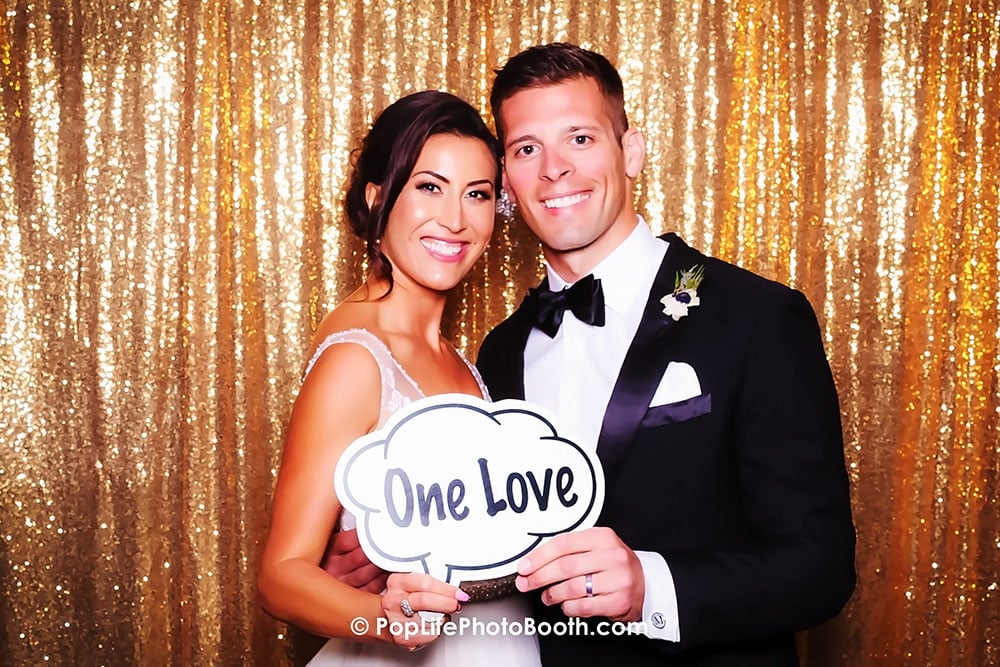 Happy newly married couple holding a sign that says "One Love" in a photo booth with a sparkling gold backdrop, capturing joyful wedding memories.