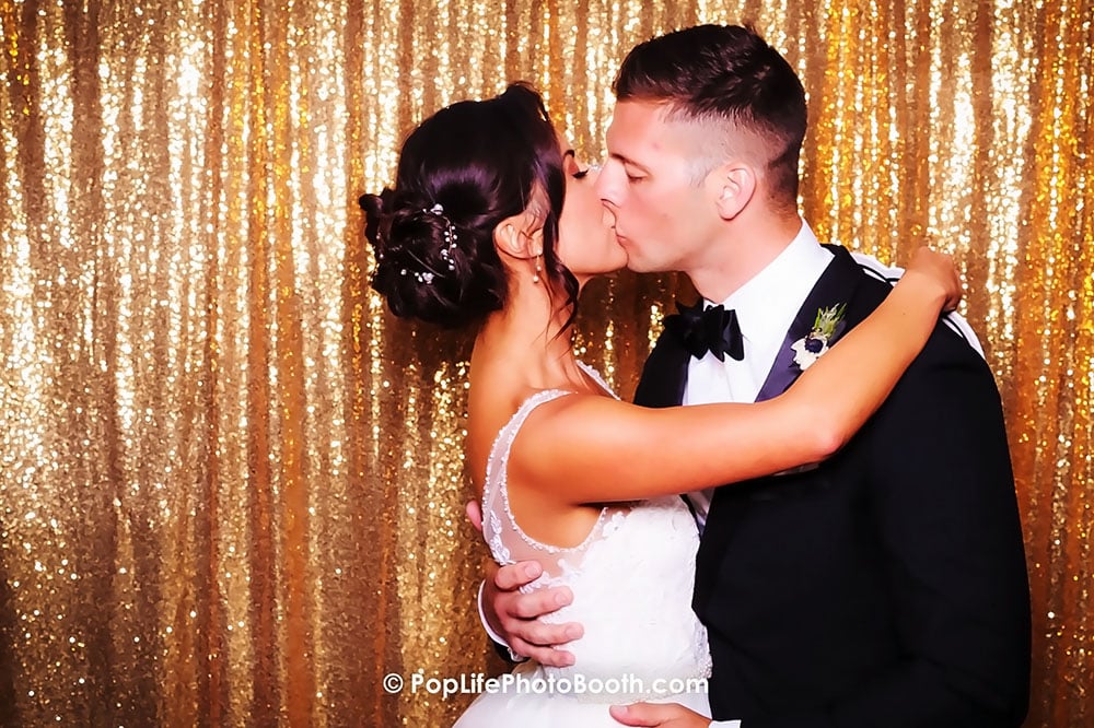 Couple kissing in front of a shimmering gold backdrop, celebrating their wedding with joy and love, capturing memorable moments with Pop Life Photo's photo booth.