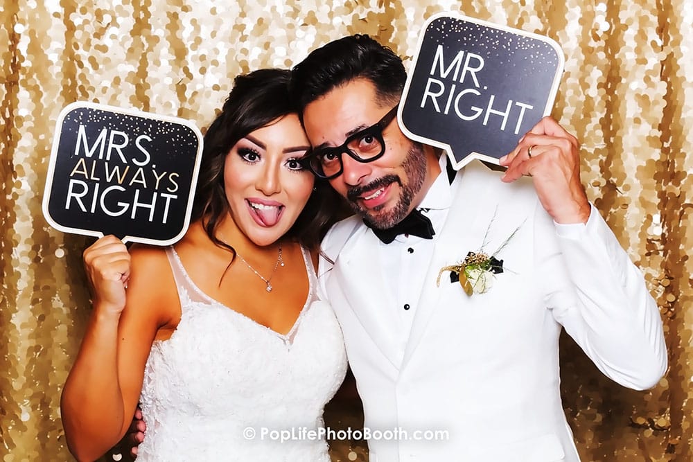 Happy couple at a wedding photo booth holding signs that say "MRS. ALWAYS RIGHT" and "MR. RIGHT," with a sparkling gold backdrop.