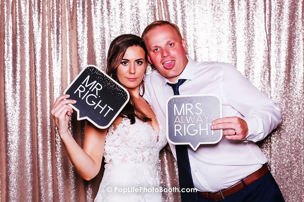 Happy newly married couple posing in photo booth, holding signs "MR. RIGHT" and "MRS. ALWAYS RIGHT," with a sparkling backdrop, capturing joyful wedding memories.