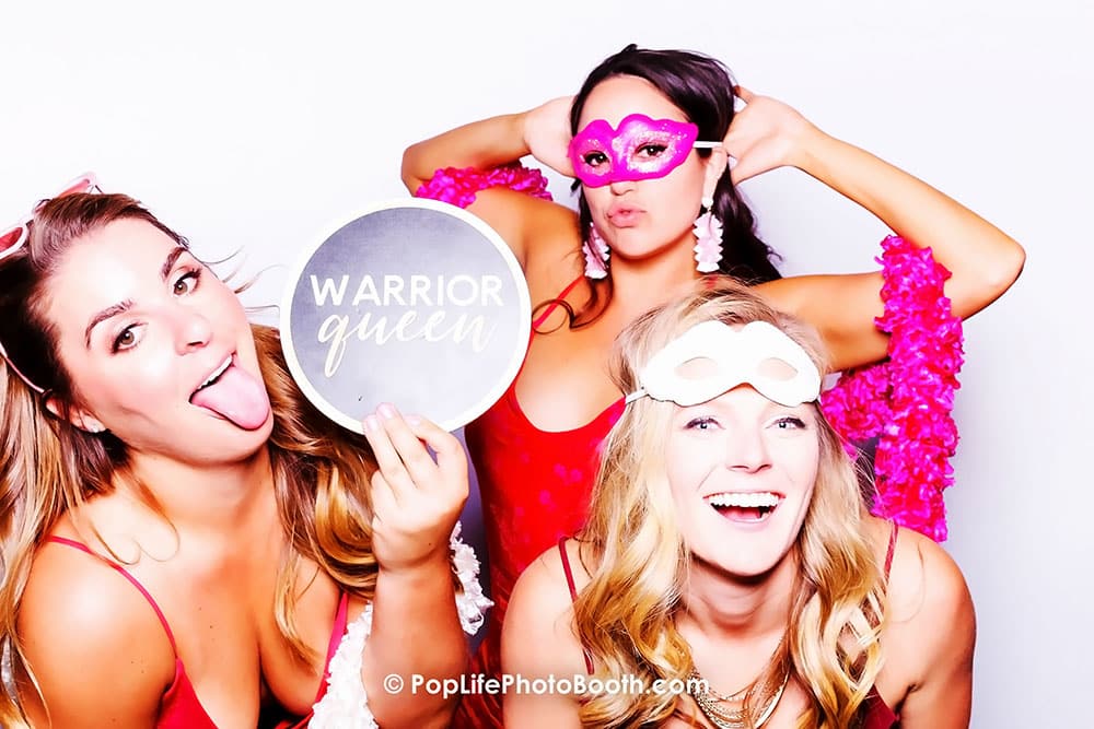 Three women in vibrant outfits posing joyfully in a photo booth, one holding a sign reading "WARRIOR QUEEN," showcasing fun and celebration at a wedding event.