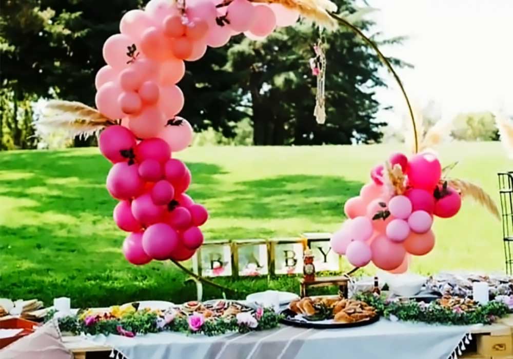 Boho picnic-style setup featuring a pink balloon arch and decorative table with food, flowers, and "BABY" sign, ideal for intimate outdoor events.