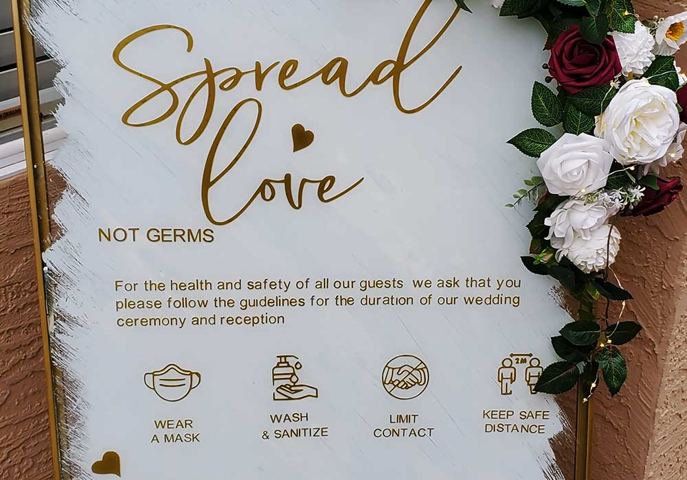 Covid-19 wedding safety sign with "Spread love, not germs" message, featuring guidelines to wear masks, wash hands, limit contact, and maintain social distance, decorated with flowers.