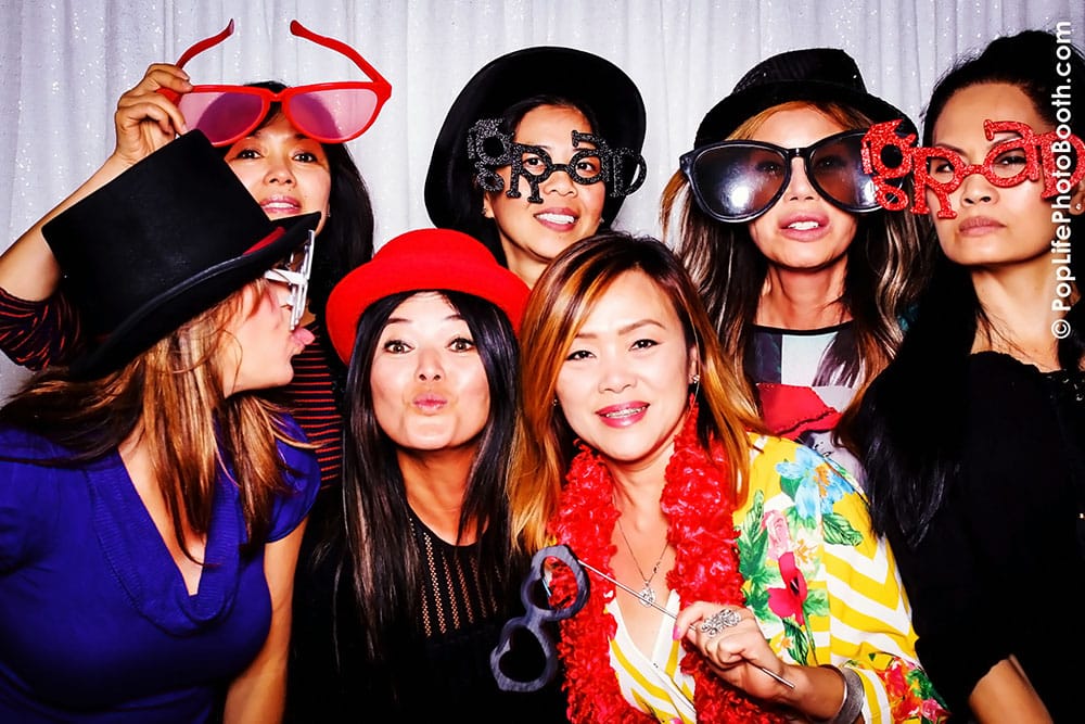 Group of women wearing fun hats and oversized glasses posing playfully in a photo booth, capturing joyful moments at a private event.