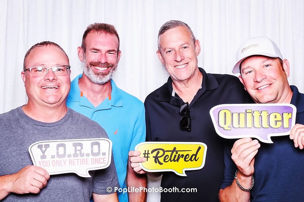 Four men smiling in a photo booth, holding signs that read "Y.O.R.O. YOU ONLY RETIRE ONCE," "#Retired," and "Quitter," capturing joyful moments at a private event.