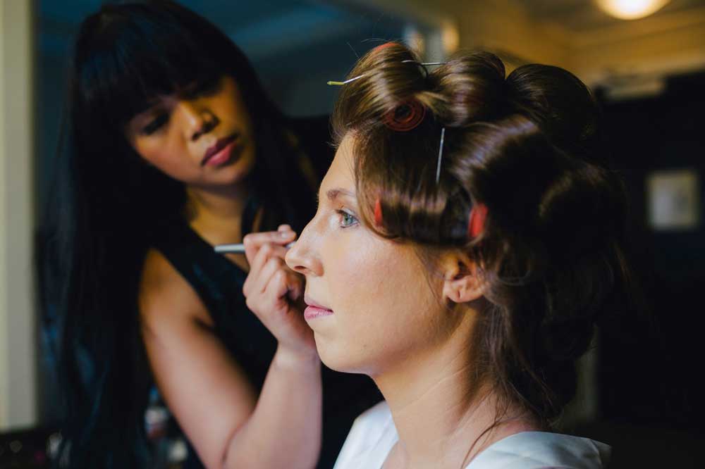 Bride receiving hairstyling and makeup application from a professional artist, featuring classic curls and a serene expression, in a wedding preparation setting.