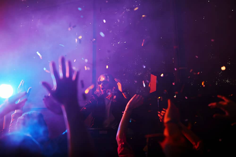 Modern DJ performing at turntables, creating an energetic atmosphere with colorful lighting and confetti, surrounded by enthusiastic audience hands in a smoky nightclub setting.