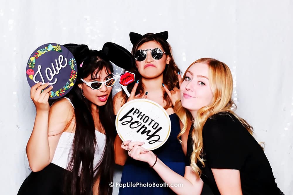 Three women posing playfully in a photo booth, holding fun props including signs that say "Love" and "Photo Bomb," with a festive backdrop, capturing the joyful spirit of wedding celebrations.