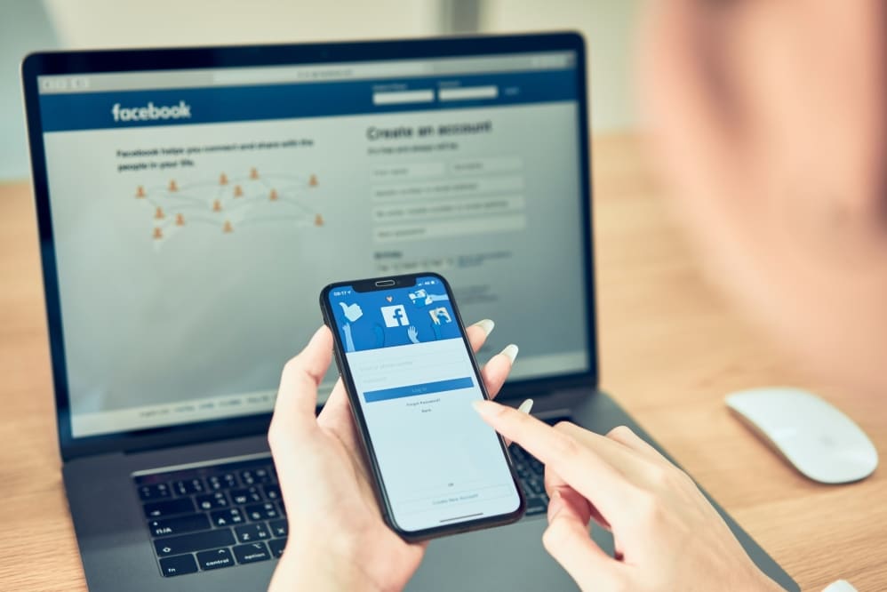 Woman holding smartphone, accessing Facebook login screen, with laptop displaying Facebook homepage, emphasizing social media engagement for event promotion.