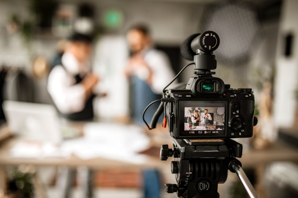 Camera on tripod capturing live video of event hosts interacting, with promotional materials in a modern workspace setting.