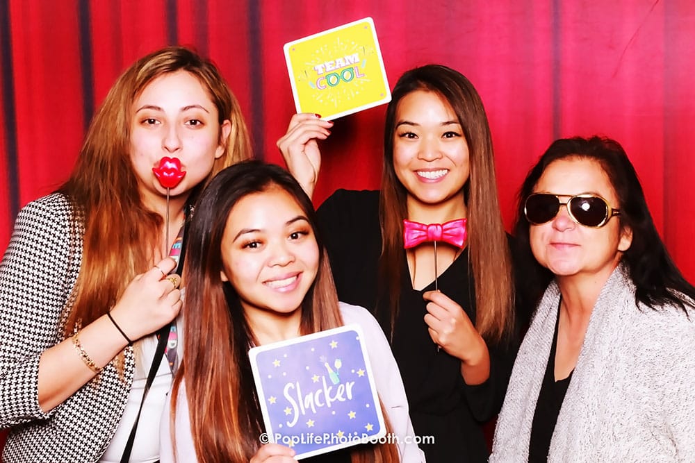 Four women posing in a photo booth, holding fun props like a rose and signs reading "Slacker" and "Totally Cool," against a vibrant red backdrop, capturing the lively spirit of corporate event entertainment.