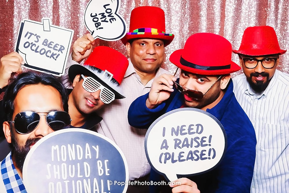 Group of men wearing red hats and sunglasses in a photo booth, holding signs with playful messages like "It's Beer O'Clock" and "I Need a Raise! (Please!)", creating a fun atmosphere at a corporate event.