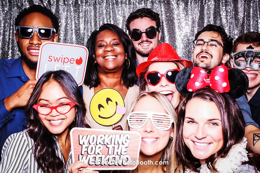 Group of smiling individuals posing in a photo booth with fun props, including signs that say "Swipe Left" and "Working for the Weekend," showcasing a lively atmosphere ideal for corporate events and enhancing brand visibility.
