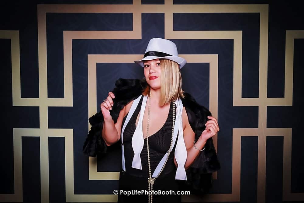 Woman in stylish costume posing in a photo booth at a corporate event, wearing a hat and feather boa, with a decorative geometric backdrop.