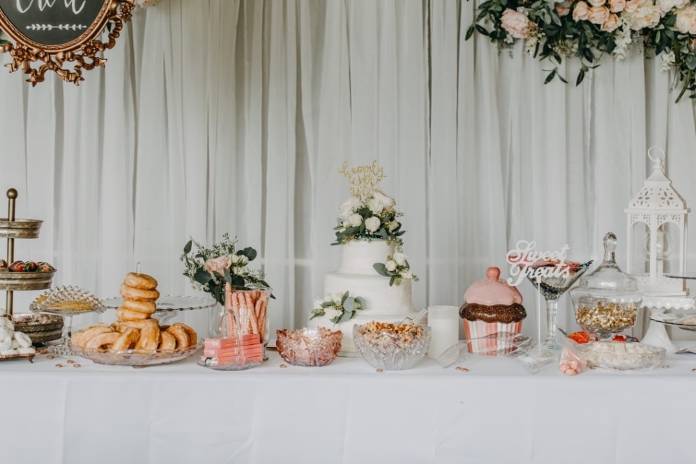 Wedding dessert table featuring a multi-tiered cake, assorted sweet treats like donuts and chocolate-covered snacks, floral arrangements, and decorative signage reading "Sweet Treats."