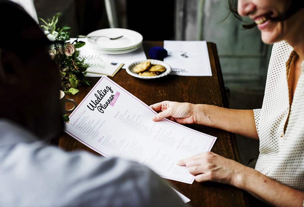 Couple reviewing a wedding planner checklist at a table with snacks, emphasizing wedding planning and organization for a successful event.