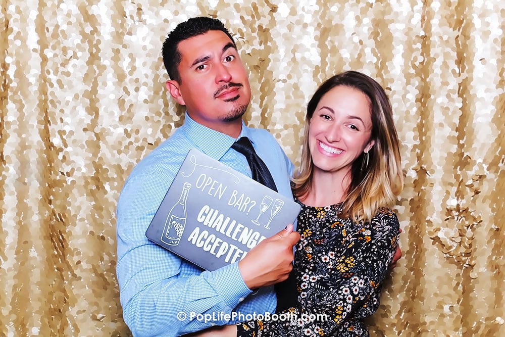 Couple posing in a photo booth with a sign reading "Open Bar - Challenge Accepted," against a shimmering gold backdrop, highlighting fun and entertainment at a wedding celebration.