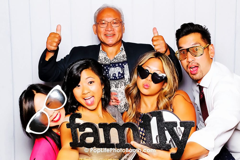 Group of five people having fun in a photo booth, holding a "family" sign, showcasing playful expressions and accessories, ideal for family celebrations at weddings.