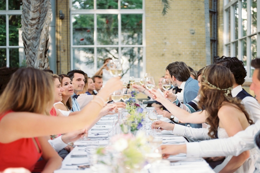 Guests toasting at a wedding reception, celebrating with drinks at a long table adorned with flowers, in a bright, airy venue.