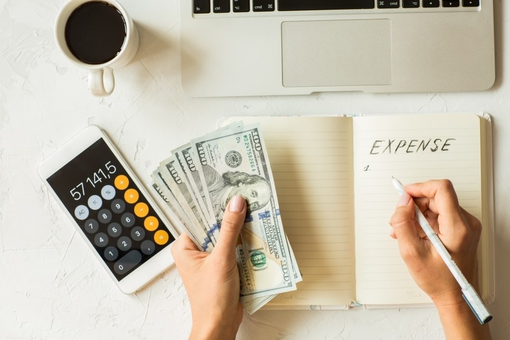 Hand holding cash and writing in a notebook labeled "EXPENSE," with a calculator displaying a total, alongside a laptop and coffee cup, illustrating wedding budget tracking and expense management.