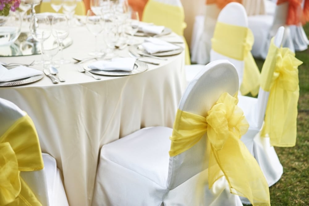Wedding reception table setup with muted peach and yellow accents, featuring elegantly arranged chairs with yellow sashes, elegant tableware, and empty glasses, reflecting a spring wedding theme.