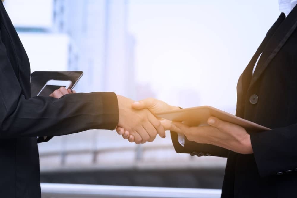 Business professionals shaking hands to symbolize partnership and commitment, with tablets in hand, set against a modern office backdrop.