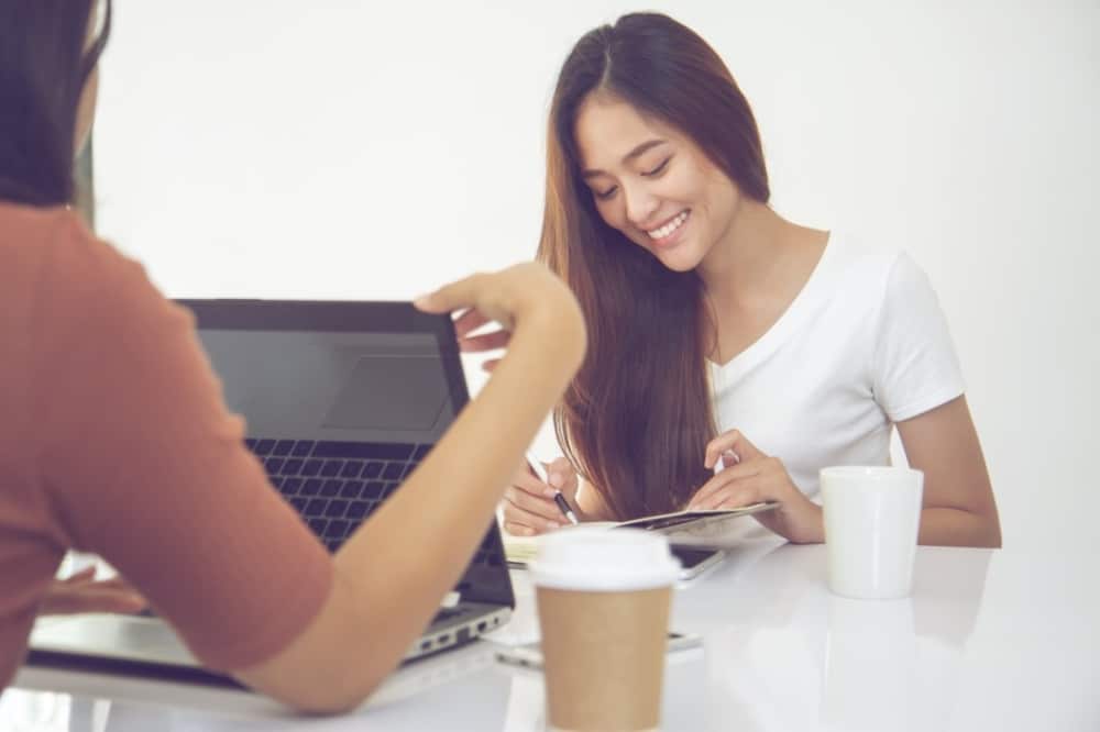 Young woman smiling while studying and discussing plans with a friend over coffee and laptop, emphasizing collaboration in event planning.