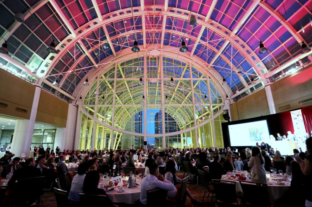 Elegant fundraising event inside a spacious venue with a high glass ceiling, featuring numerous tables filled with guests, illuminated by colorful lights and a large presentation screen.