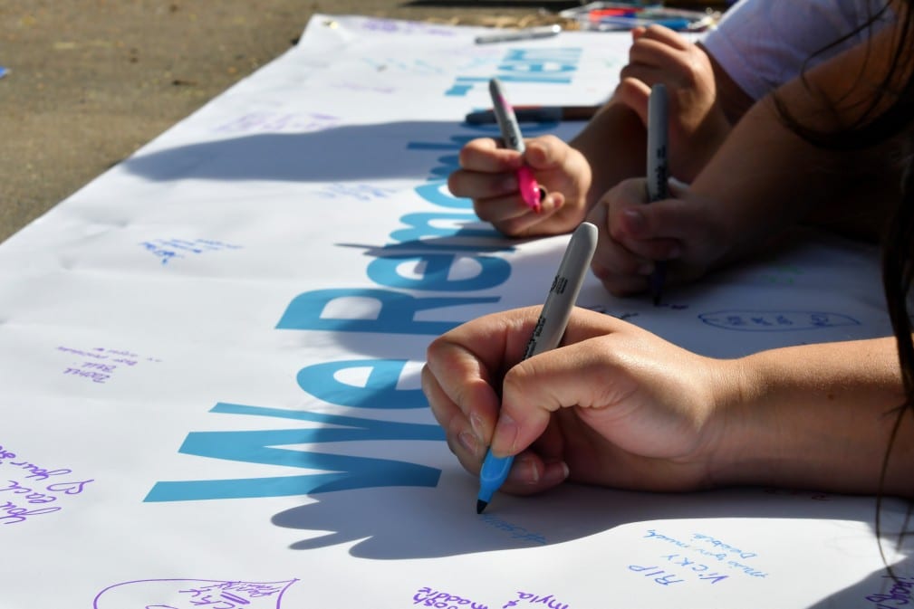 Hands writing messages on a remembrance banner during a fundraiser event for mental health awareness, featuring the phrase "We Remember" prominently displayed.